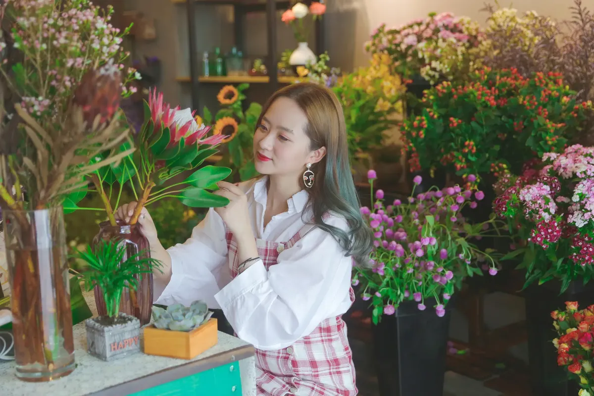 Young smiling ethnic woman in trendy wear with earring enjoying bright blooming flower with leaves in transparent vase while sitting at table in shop