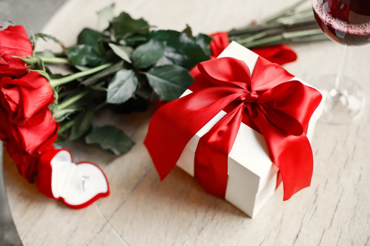 A close-up of a romantic gift box adorned with a red ribbon, beside red roses and a glass of red wine.